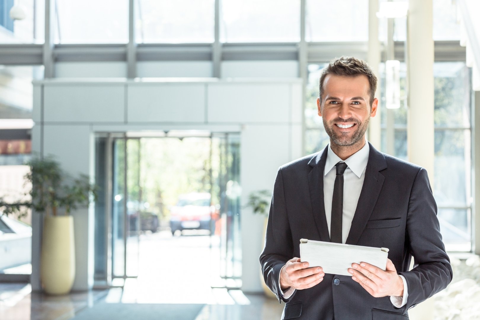 Smiling hotel manager in a suit holding a digital tablet in a bright, modern hotel lobby