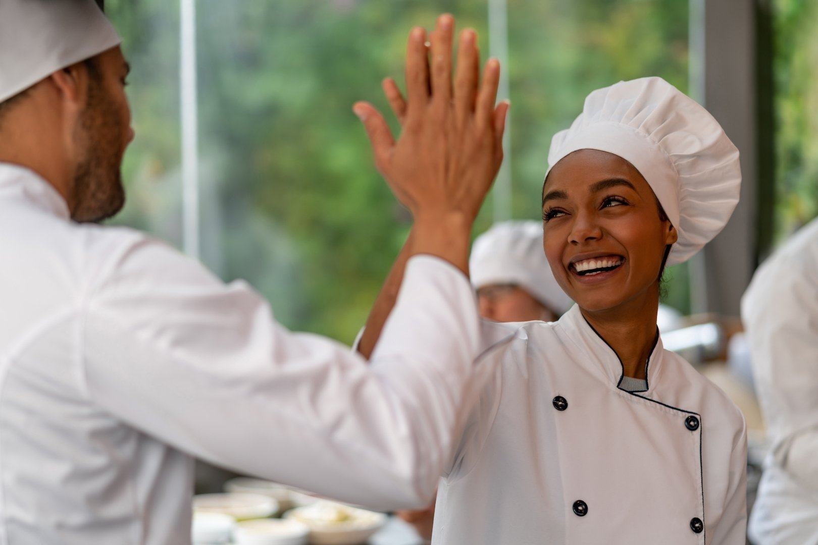 Two smiling chefs in uniform high-fiving in a kitchen, celebrating teamwork and a positive work environment.