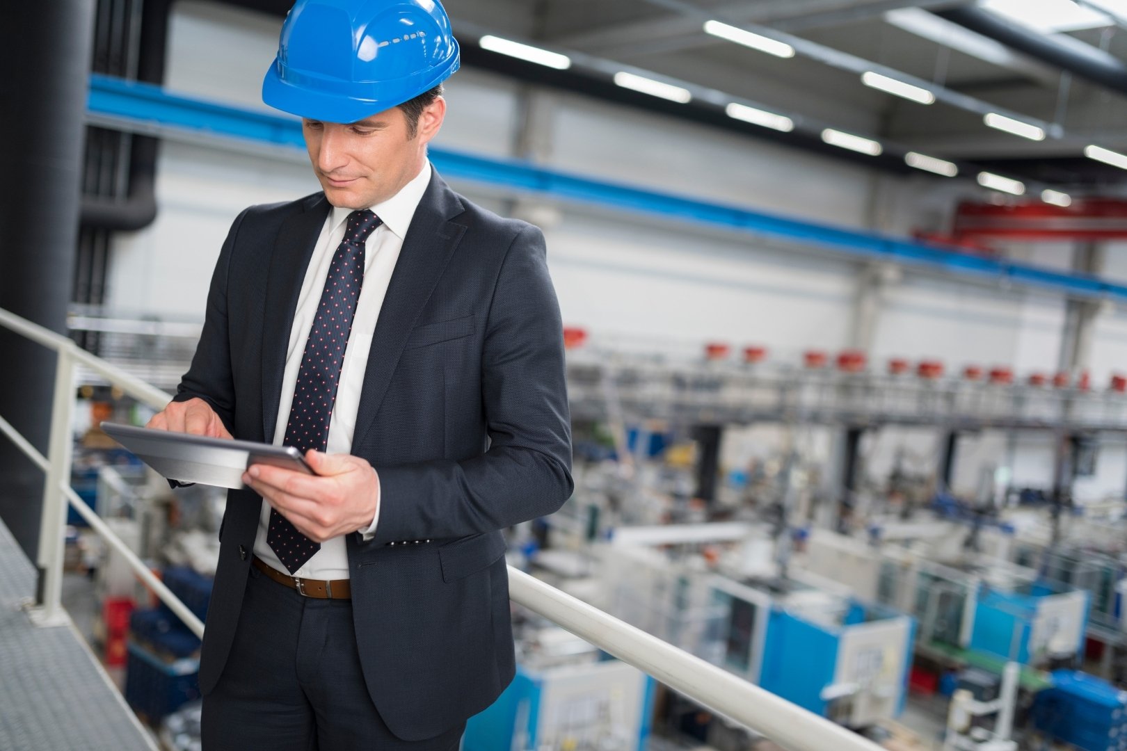 Manufacturing manager in a suit and blue hard hat using a tablet while overseeing production on a factory floor.
